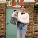 Une jeune femme souriante devant une porte bleue, portant le sac de transport gris de la marque Kavee avec un cochon d'inde dedans.